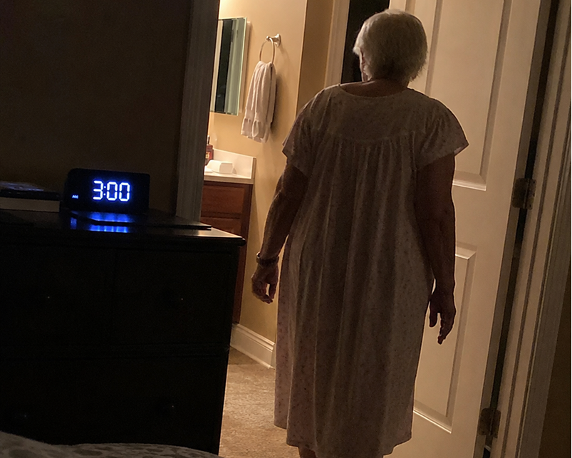 An older woman viewed from behind wearing a light beige patterned nightgown in a dimly lit bedroom, standing near an open door next to a dark dresser with a blue digital alarm clock showing 3:00.