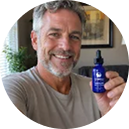 A man holding sleepwhale dropper bottle, shown inside a circular frame. The background is a warm beige indoor setting with soft-focus greenery.
