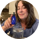 A woman holding sleepwhale bottle in front of a blurred kitchen background, with a glass of water in the foreground.