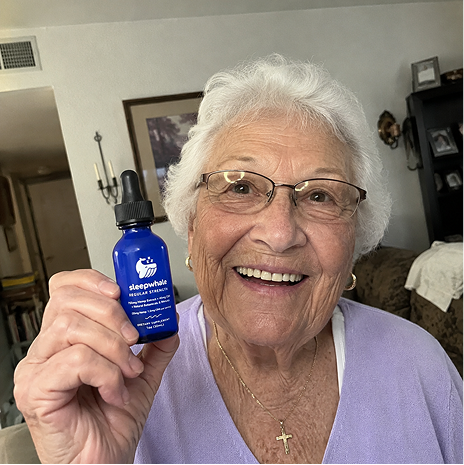 An elderly woman with white hair and glasses smiles while holding up a small blue dropper bottle labeled 'blue powerhouse' in a home setting.